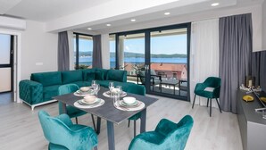 A modern living room with a teal sofa, dining table set for four, and large windows offering a view of the sea.