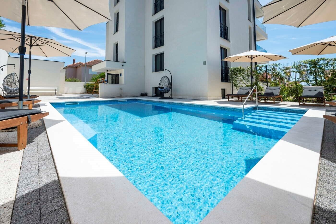A rectangular swimming pool with clear blue water, surrounded by lounge chairs and umbrellas, set against a modern building facade.