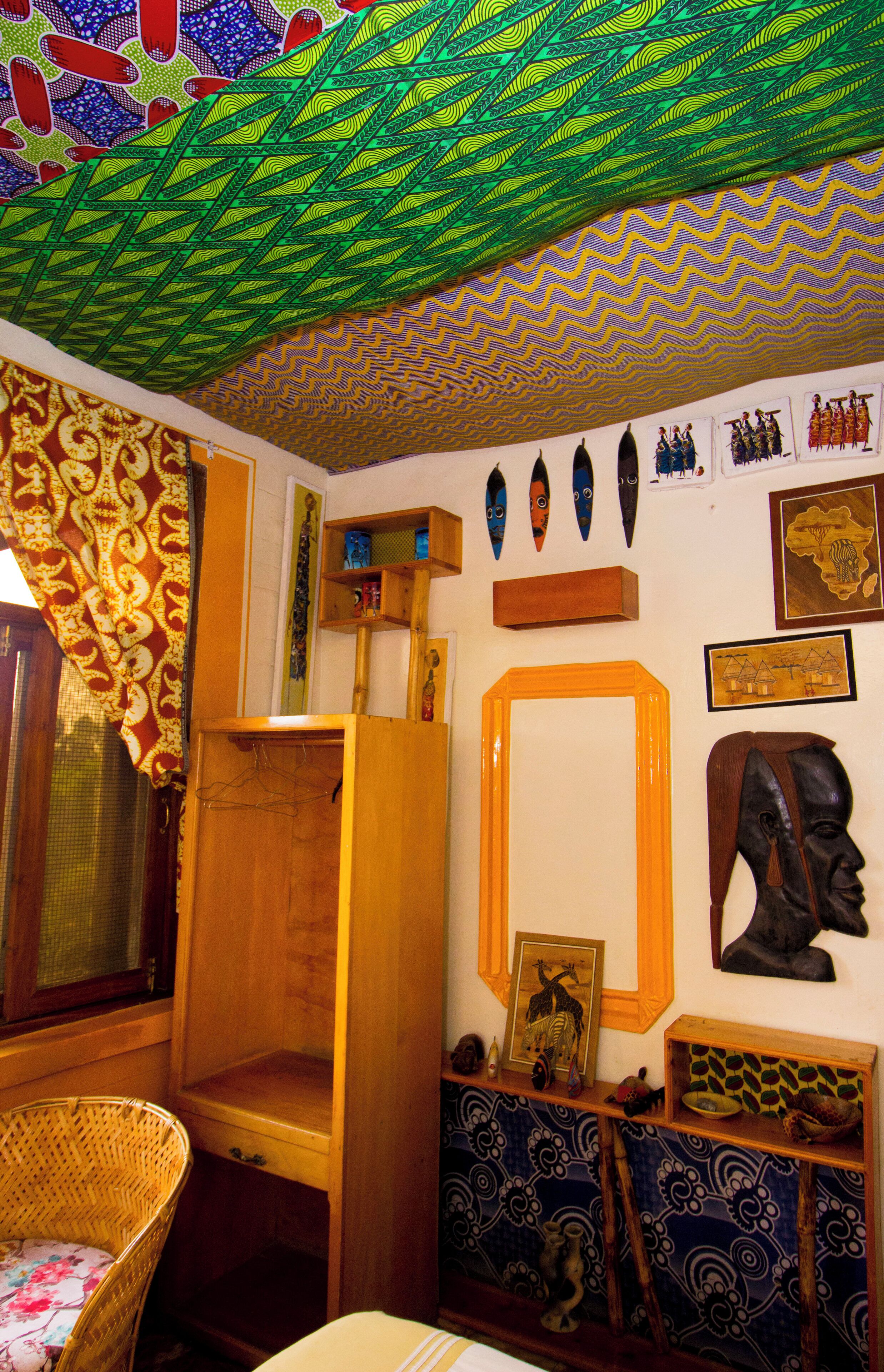 A room with a patterned ceiling, a wooden cabinet, a wicker chair, and various wall decorations including masks and framed pictures.