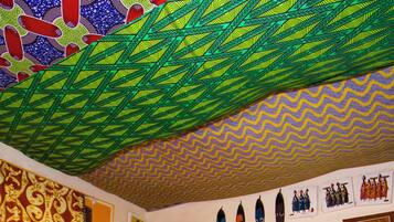 A room with a patterned ceiling, a wooden cabinet, a wicker chair, and various wall decorations including masks and framed pictures.