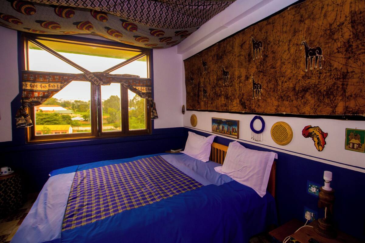 Een slaapkamer met een groot bed, een raam met gordijnen, een muur met tribale kunst en uitzicht op groen.