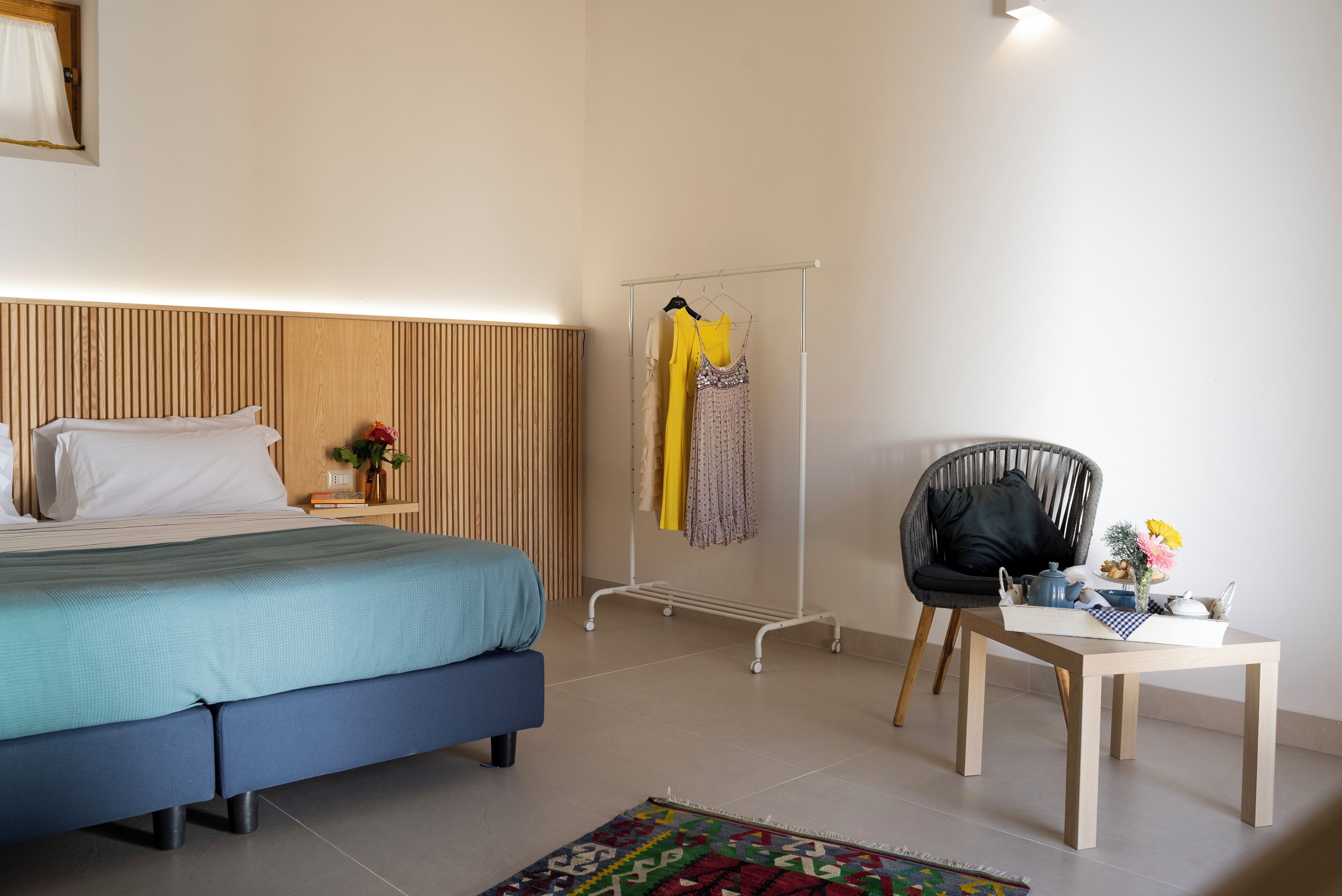 Un dormitorio con cama, mesita de noche con lámpara, una silla y una pequeña mesa con un jarrón de flores.