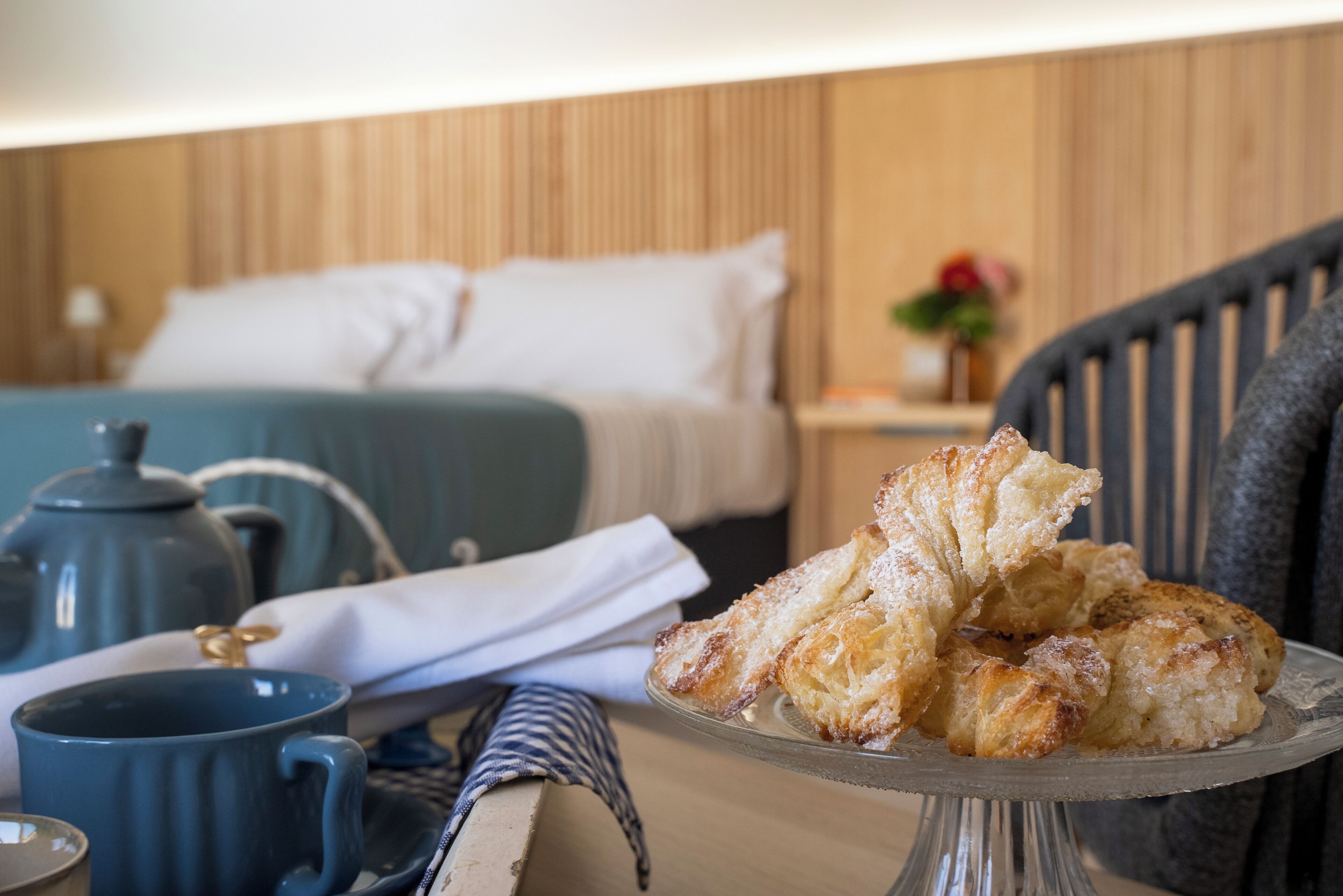 A neatly made bed with white linens, a blue mug, a teapot, and a plate of pastries on a glass stand.