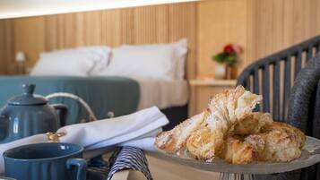 A neatly made bed with white linens, a blue mug, a teapot, and a plate of pastries on a glass stand.