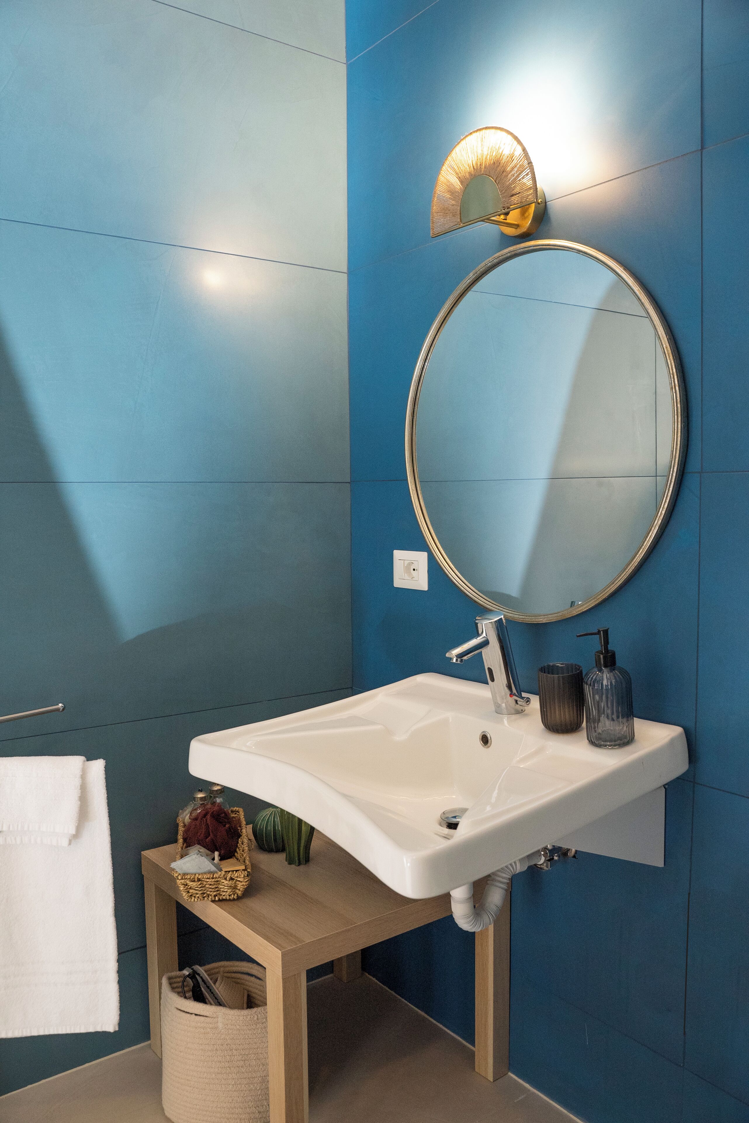 A bathroom with a white sink, a round mirror, a wall-mounted light, and a towel hanging on the wall.