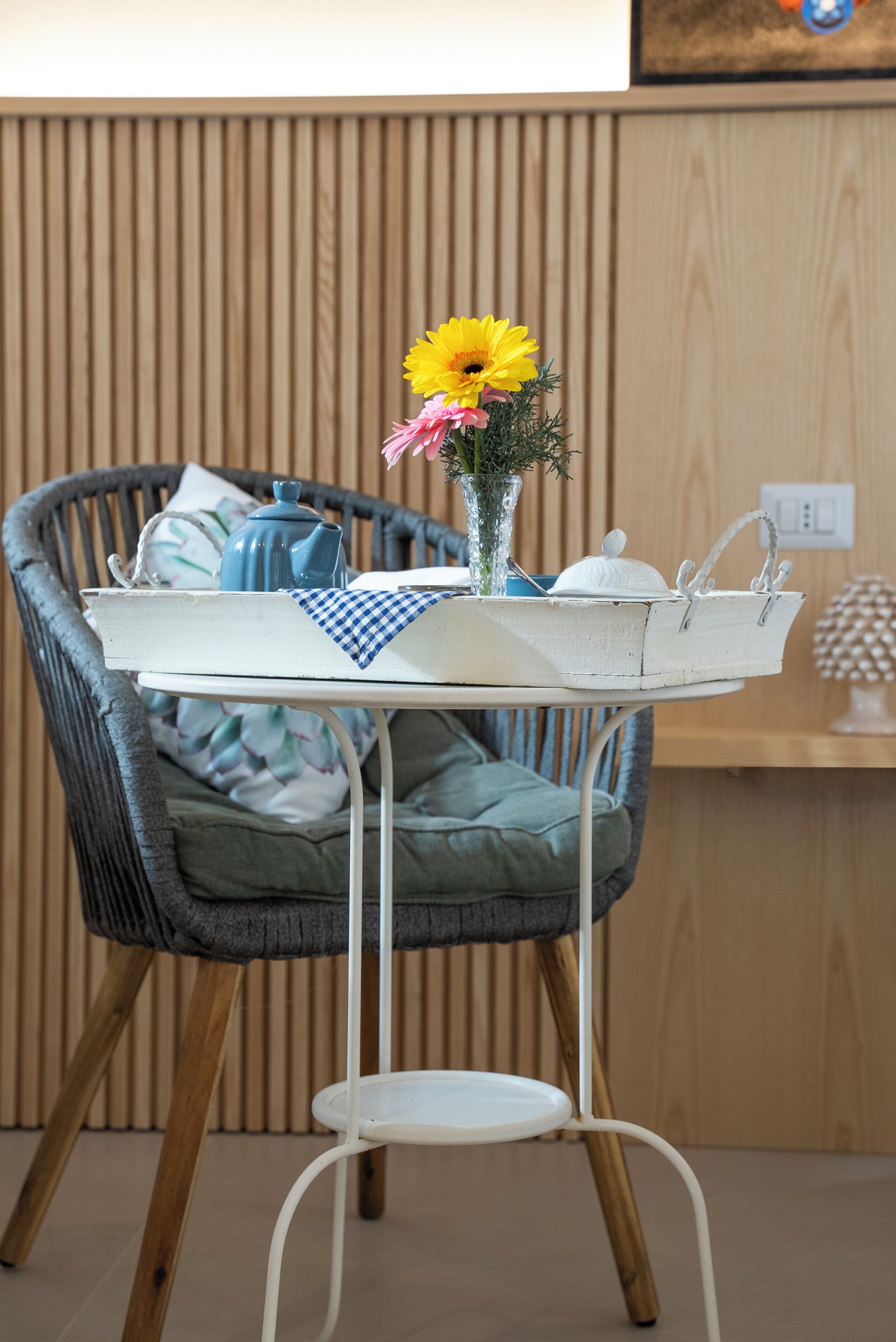 Una silla de mimbre con una mesita metálica blanca adelante, un jarrón con flores y una lámpara blanca pequeña sobre la mesa.