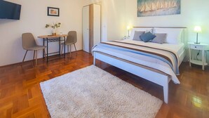 Un dormitorio con cama, mesitas de noche, un escritorio con sillas, un cuadro en la pared y un televisor fijado a la pared.