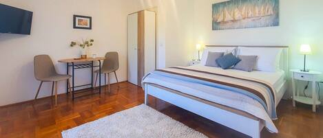 A bedroom with a bed, bedside tables, a desk with chairs, a painting on the wall, and a TV mounted on the wall.