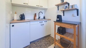 Cocina compacta con armarios blancos, una cafetera negra, un hervidor y una mesa de madera con una cesta.