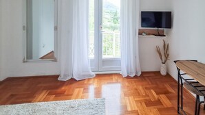 Una habitación con suelo de madera, un espejo grande, un televisor montado en la pared y un balcón con cortinas blancas.