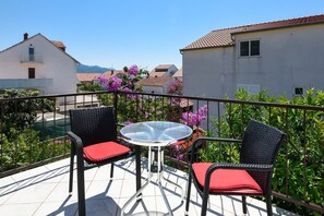 A balcony with two wicker chairs and a glass table, overlooking a garden with purple flowers and a view of houses in the distance.