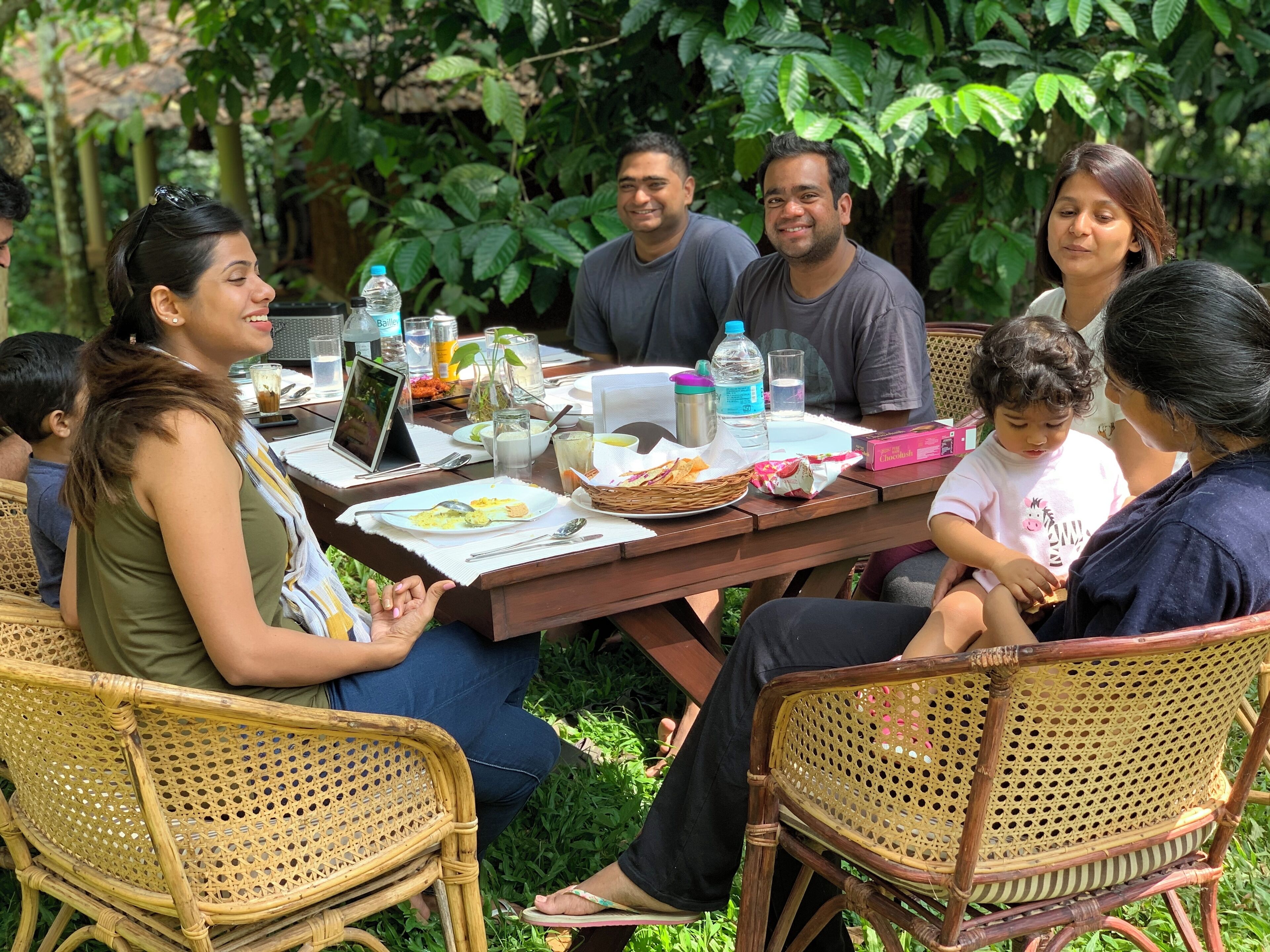 Un groupe de personnes est assis autour d’une table en bois, devant laquelle se trouvent divers objets, notamment des assiettes, des bouteilles et un ordinateur portable.