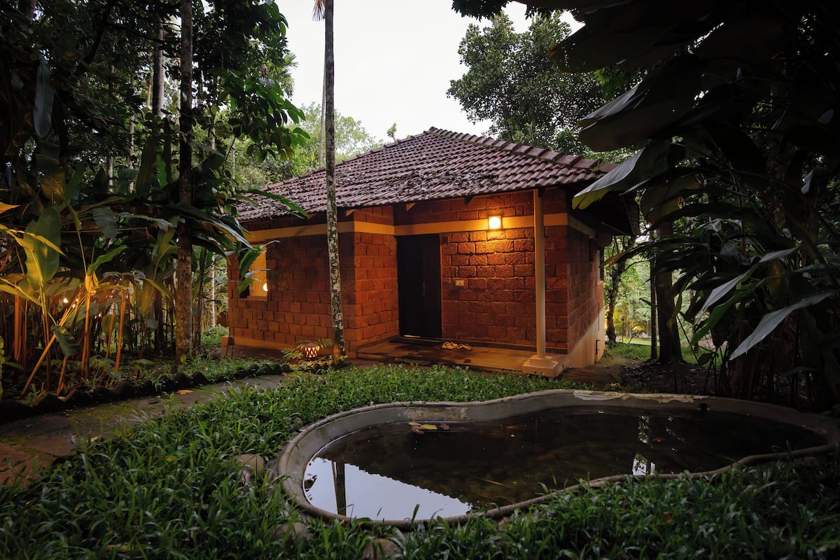 A small brick building with a tiled roof, surrounded by lush greenery and a small pond.