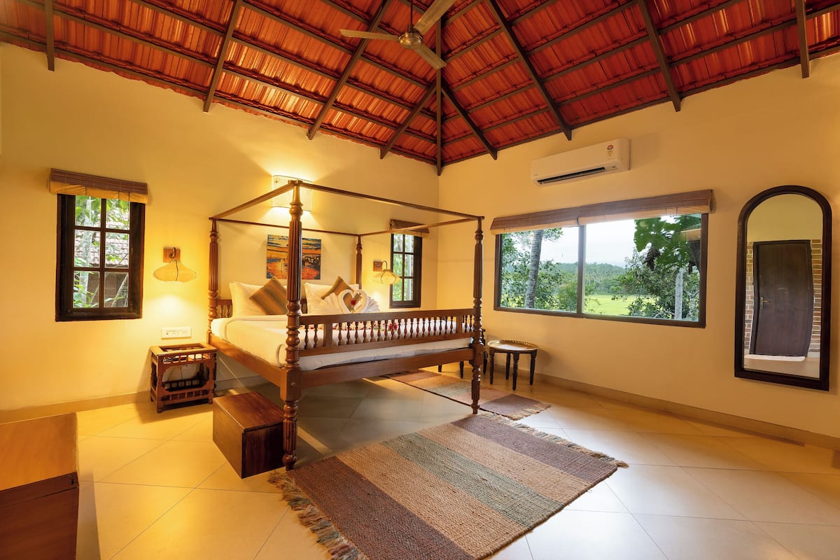 A four-poster bed with a wooden frame, a small bedside table, a wall-mounted air conditioner, and a large window with a view of greenery.