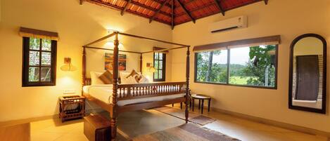 A four-poster bed with a wooden frame, a small bedside table, a wall-mounted air conditioner, and a large window with a view of greenery.