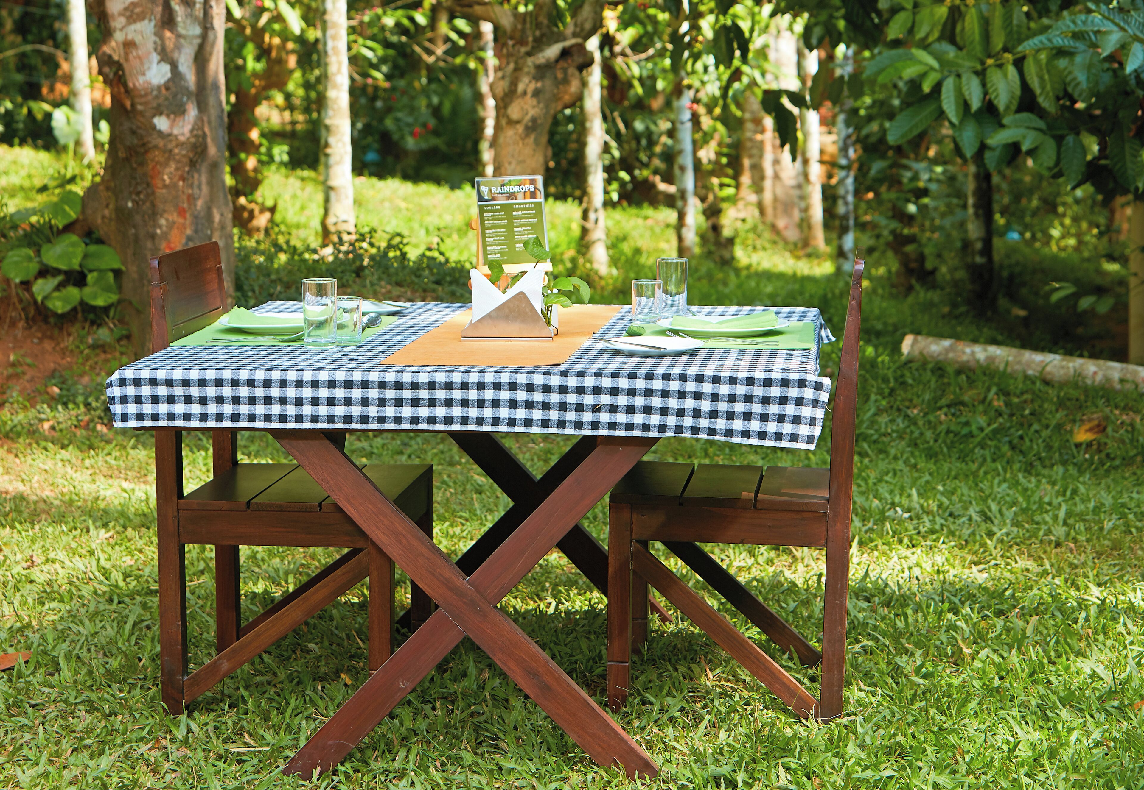 Mesa con mantel verde a cuadros, lista para usar al aire libre, con un atril para el menú y dos sillas.