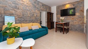 Una sala de estar con pared de piedra, un sofá color turquesa, un cojín amarillo, un armario de madera, una mesa de comedor con sillas y un televisor de pantalla plana montado en la pared.