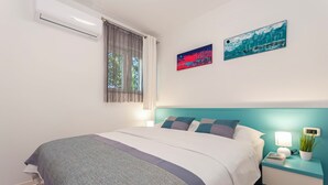 Una cama bien hecha con edredón gris y blanco, una mesita de noche azul verdoso con una lámpara y una planta en maceta, y dos obras de arte en la pared sobre la cama.