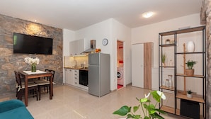 Una sala de estar moderna con una pared de acento de piedra, un televisor de pantalla plana, un comedor con mesa y sillas, una cocina con refrigerador y estufa, y una unidad de estanterías con plantas.