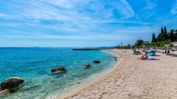 Una playa con agua cristalina, rocas y una orilla de arena. Hay sombrillas y gente en la playa.