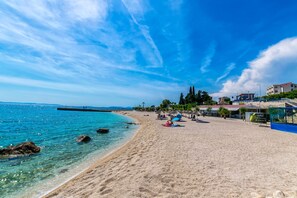 A beach with clear blue water, a sandy shore, and buildings in the background.