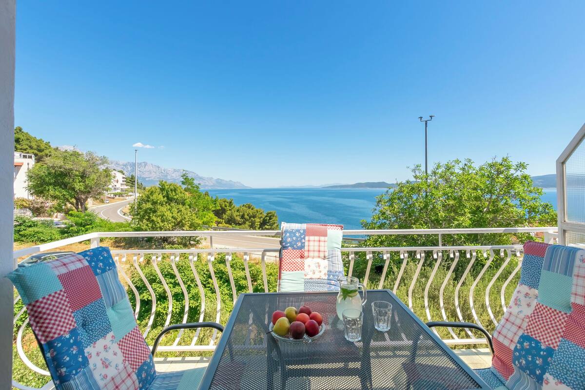 A balcony with a table set with fruits and drinks, overlooking a scenic view of a body of water and distant mountains.