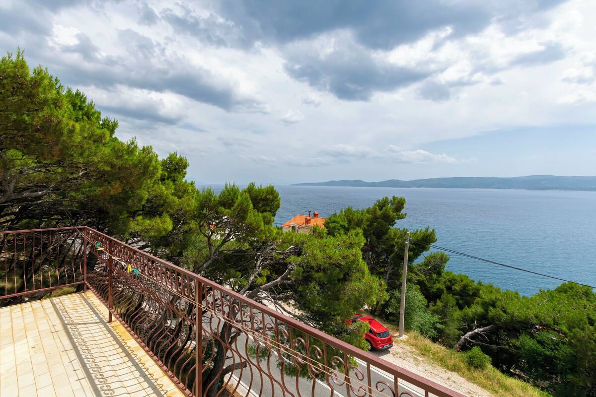 A balcony with a metal railing overlooking a coastal area with trees and a red car parked nearby.