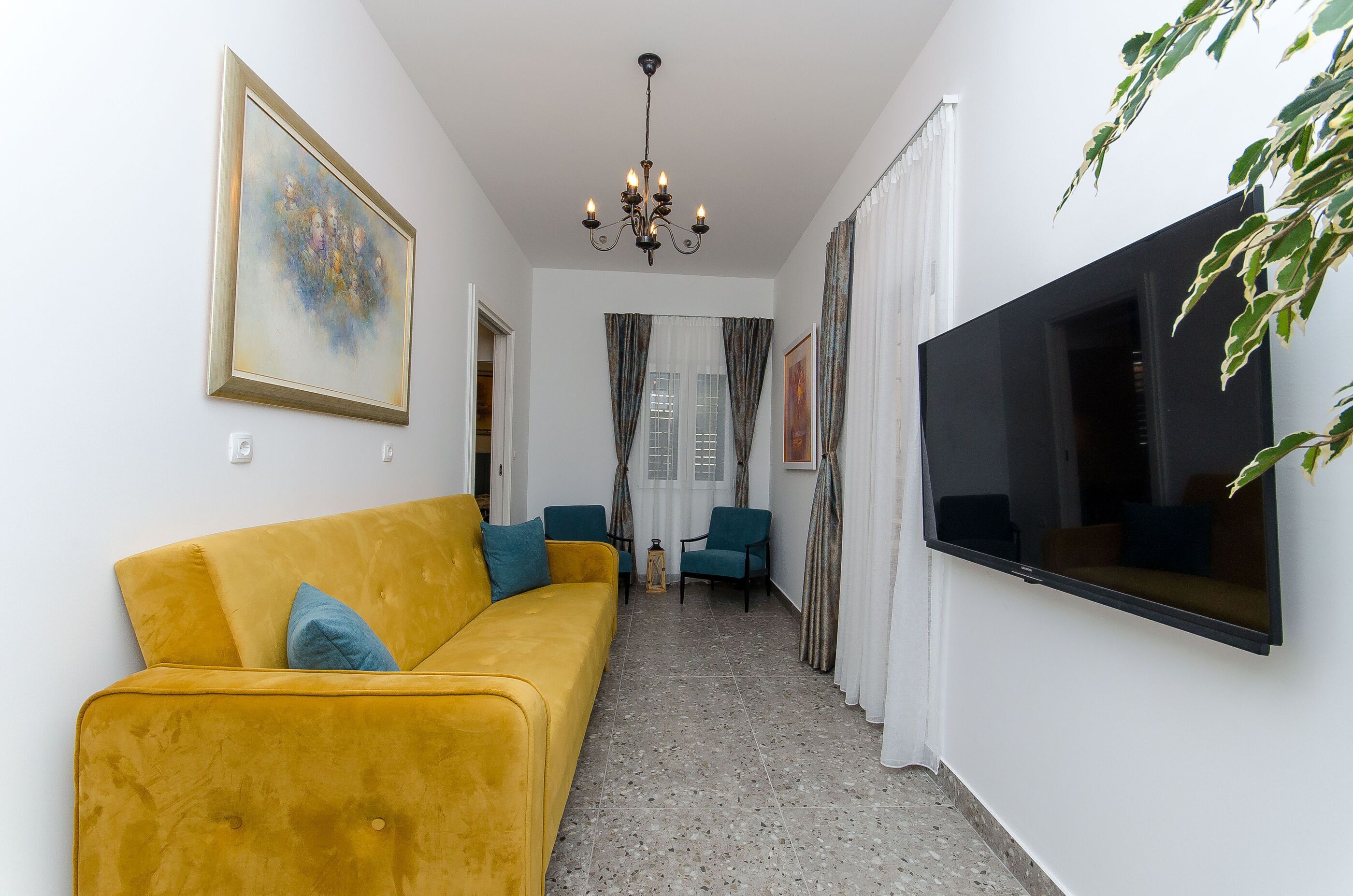 Un salon avec un canapé jaune, des fauteuils turquoise, un téléviseur et un lustre.