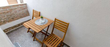 A small outdoor balcony with a wooden table and two chairs, a potted plant, and a white wall.