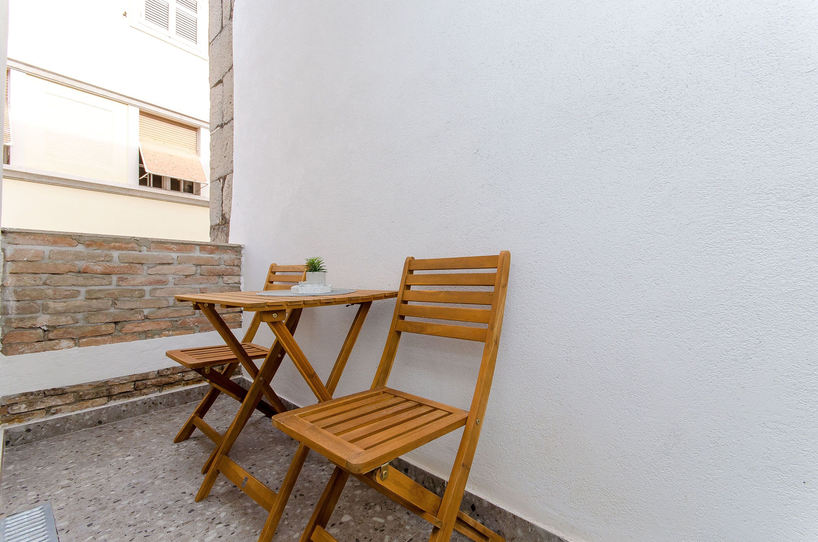 Een klein terras met een houten tafel en twee stoelen, een plant in pot en een witte muur.