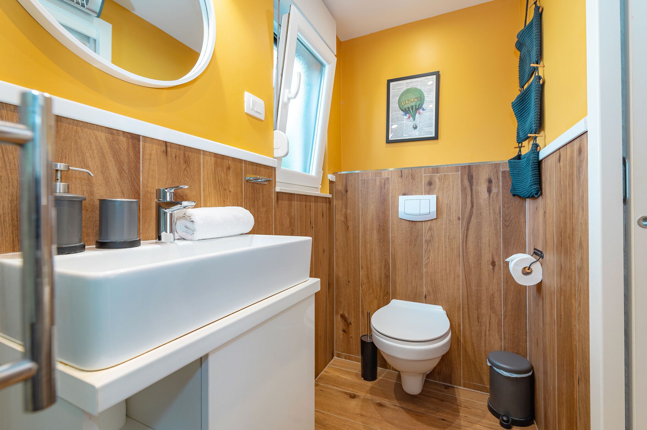 A bathroom with a wooden vanity, a white sink, a toilet, and a trash bin.