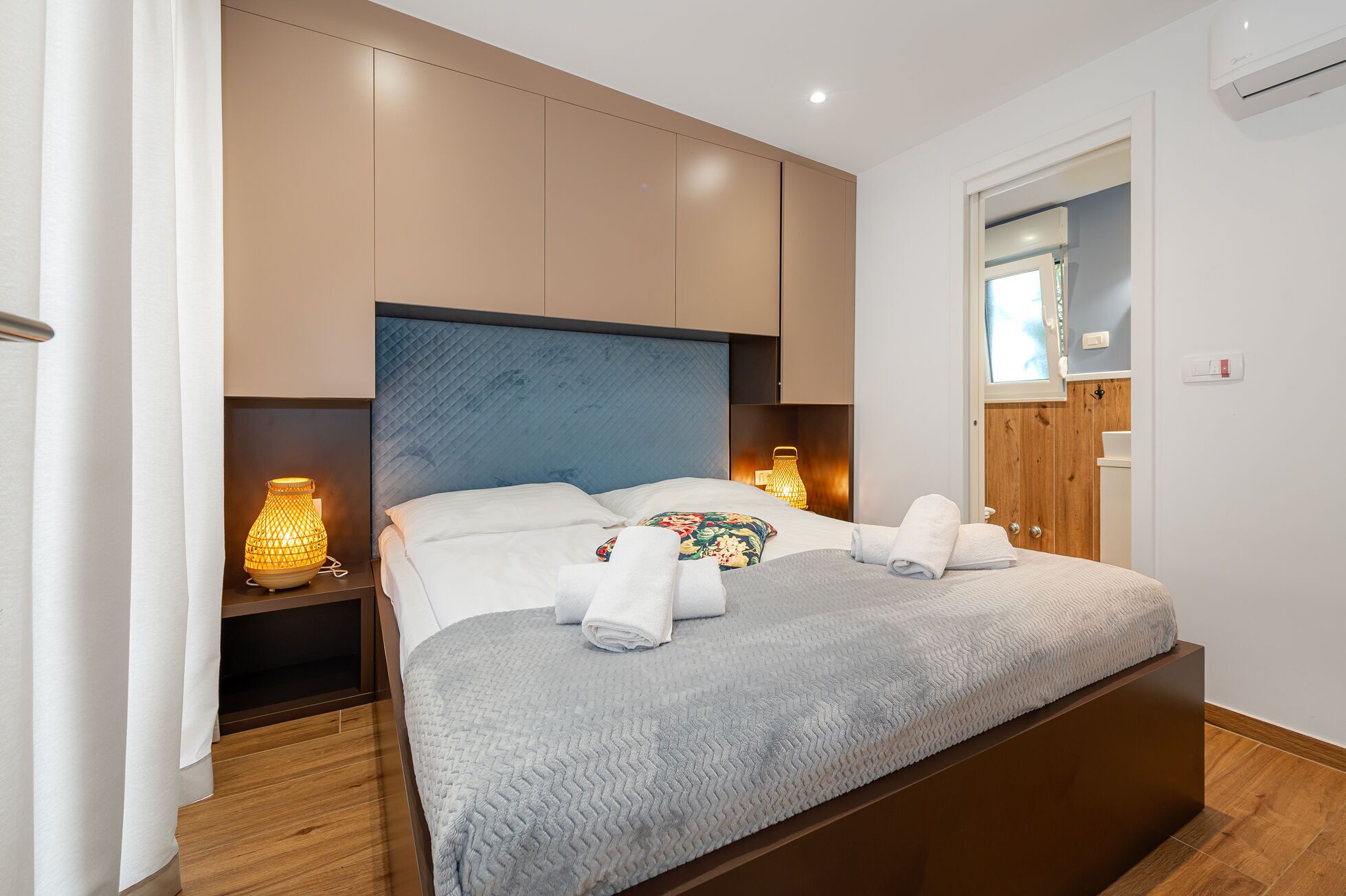 Een net opgemaakt bed met een grijze deken, twee nachtlampjes en een houten hoofdbord.
