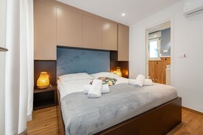 Ein ordentlich bezogenes Bett mit einer grauen Steudecke, zwei Nachttischlampen und einem hölzernen Kopfteil.