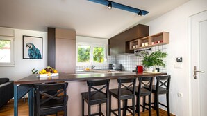 A modern kitchen with a central island, bar stools, a dining table, and a large window. - Villa Royal Green Two (Nin)