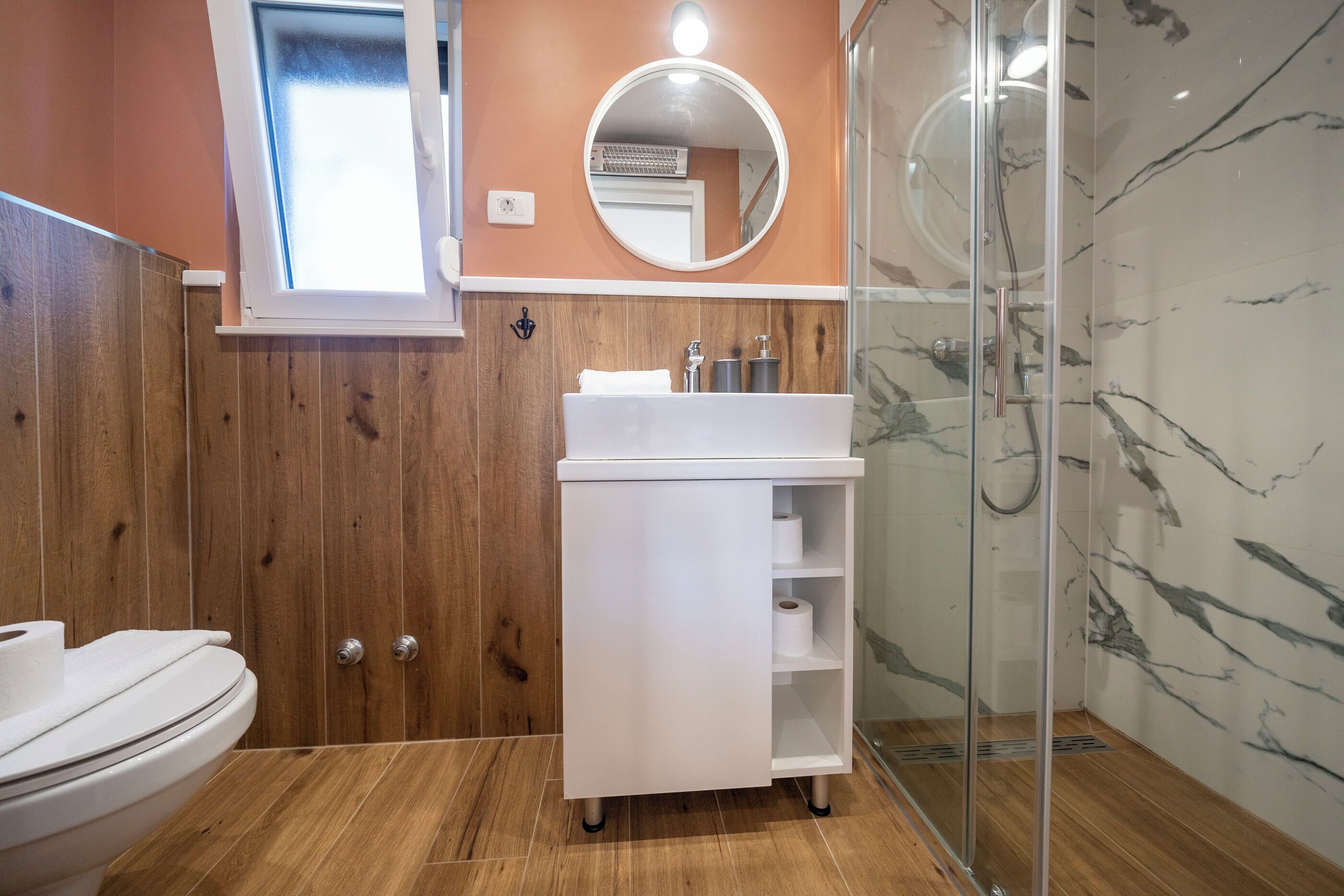 A bathroom with a wooden vanity, a toilet, a glass shower enclosure, and a marble wall.