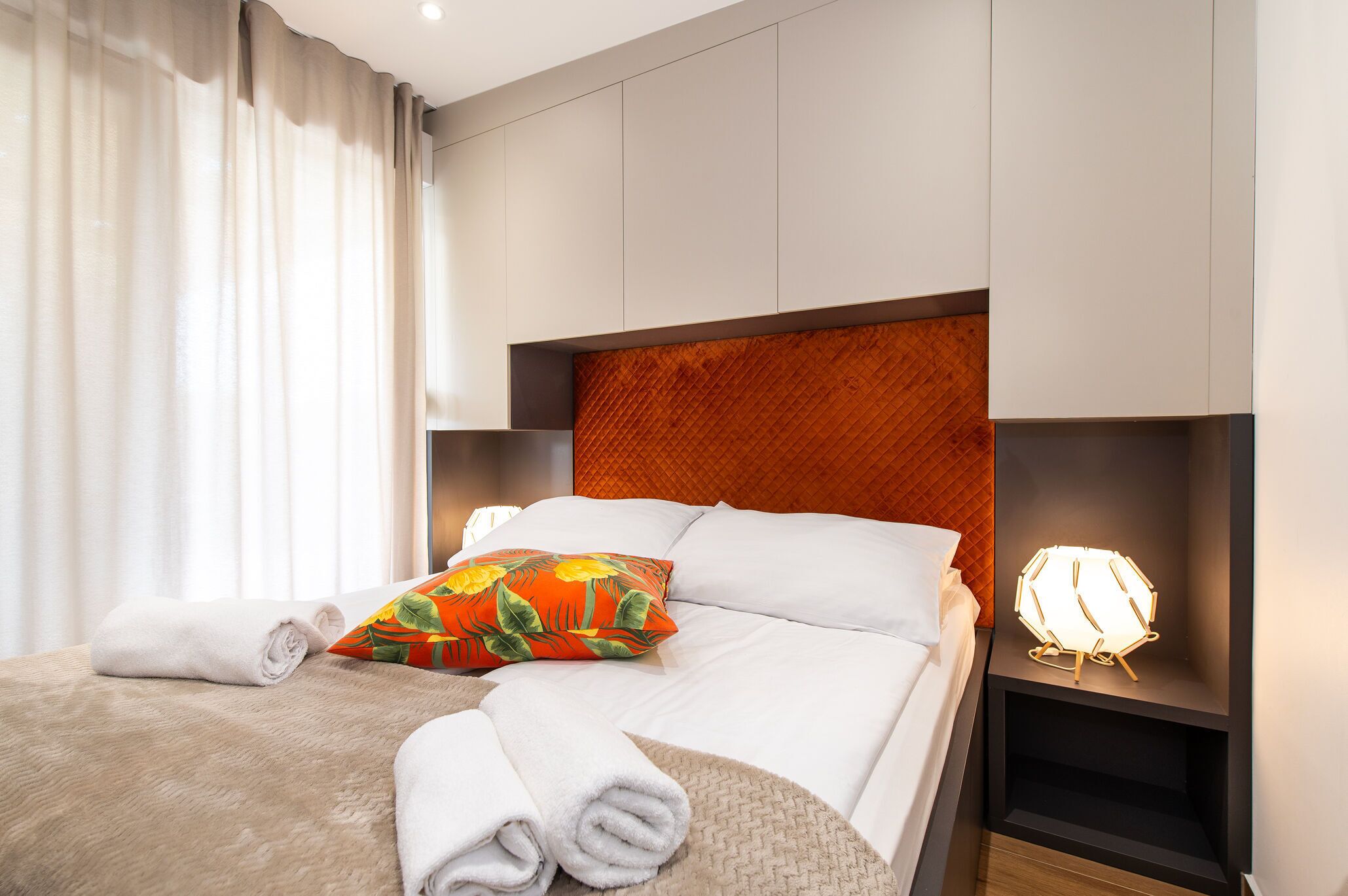 Una cama bien hecha con sábanas blancas, una almohada colorida y una lámpara de noche.