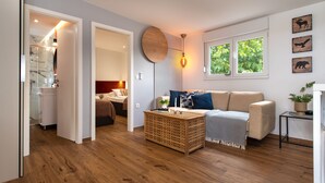 A modern living room with a wooden floor, a beige sofa, a round wall clock, and a window with a view of greenery.