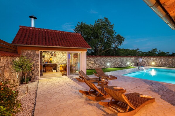 Une maison en pierre avec un toit recouvert de tuiles rouges, une piscine et des chaises longues en bois.