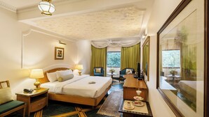 A hotel room with a bed, bedside tables, a desk, a chair, a small table with a lamp, a window with curtains, and a framed picture on the wall. - Hotel Kwality Regency (Chandigarh)