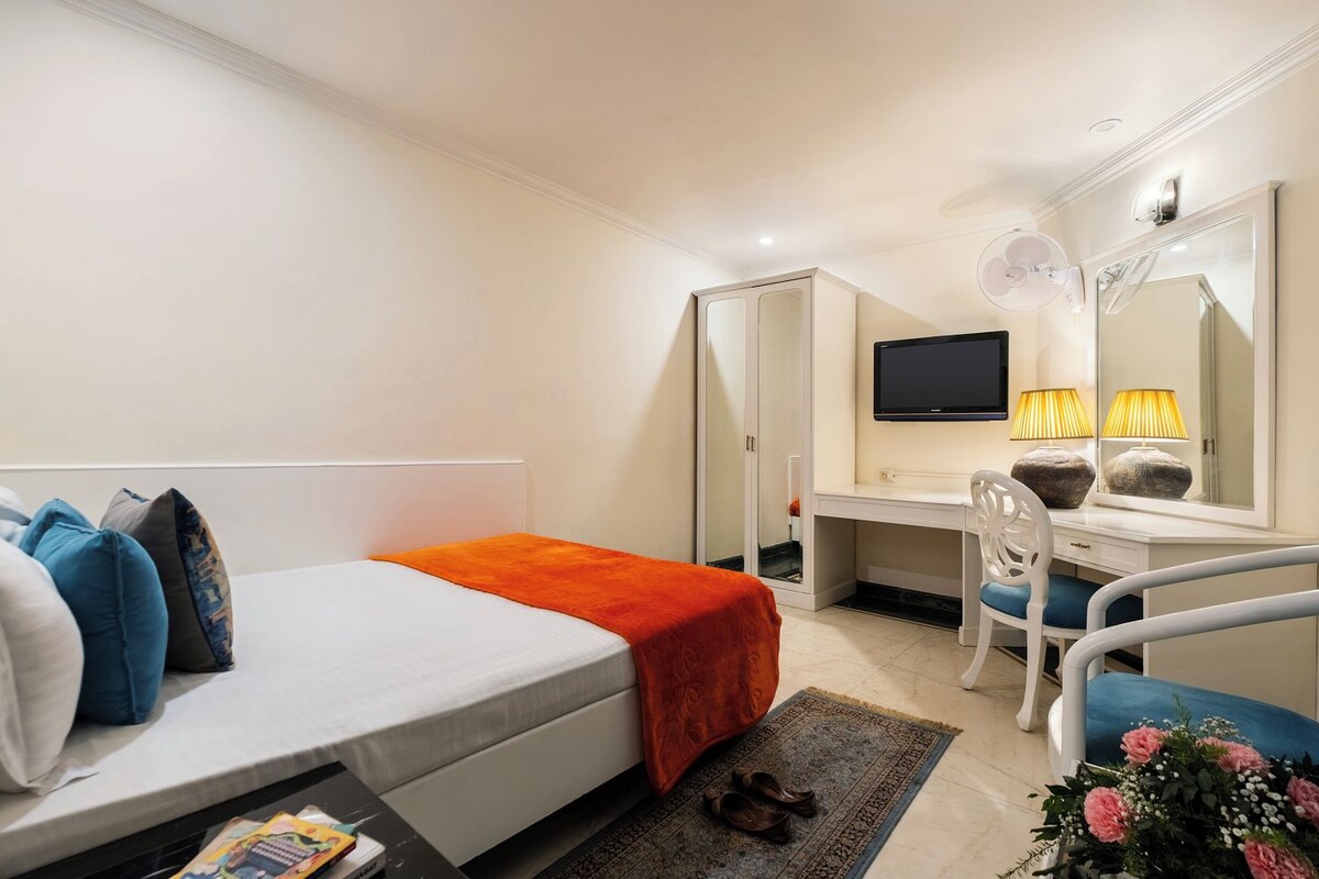 Quarto de hotel com cama, escrivaninha, cadeira, televisão e espelho.