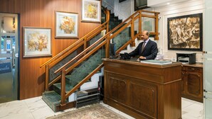 A reception desk with a man standing behind it, a wooden staircase, and framed artwork on the walls. - Hotel Kwality Regency (Chandigarh)