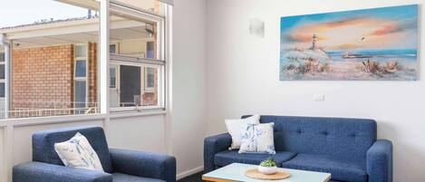 Una habitación con un sofá azul, dos sillones, una mesita con una planta, un cuadro de un faro y vistas a un edificio a través de una ventana.