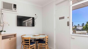 Une pièce avec un téléviseur fixé au mur, une petite table avec des chaises, un meuble en bois et une porte donnant sur la vue des bâtiments à l’extérieur.