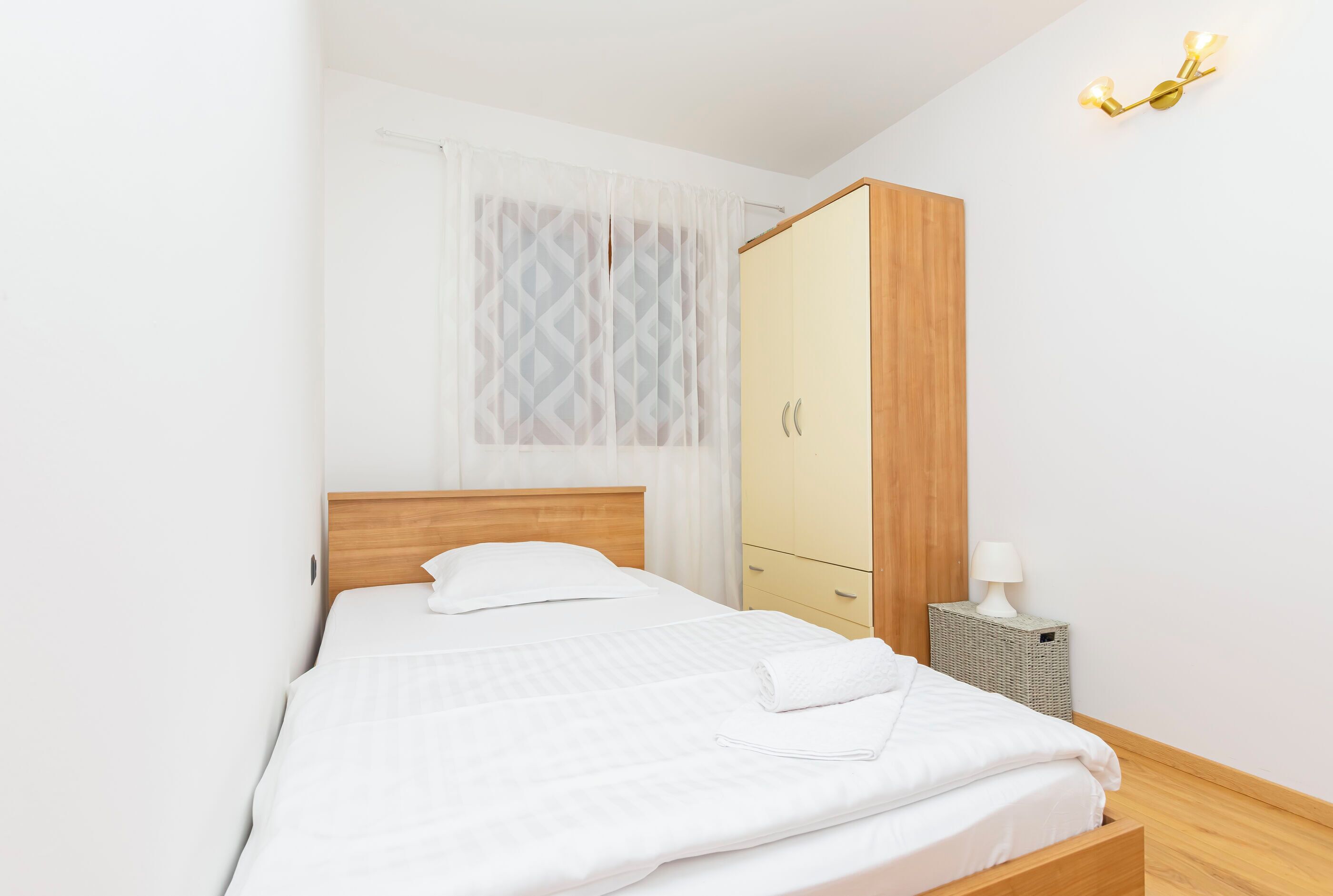 A neatly made bed with white linens, a wooden headboard, a wardrobe with a mirror, and a bedside lamp on a small table.