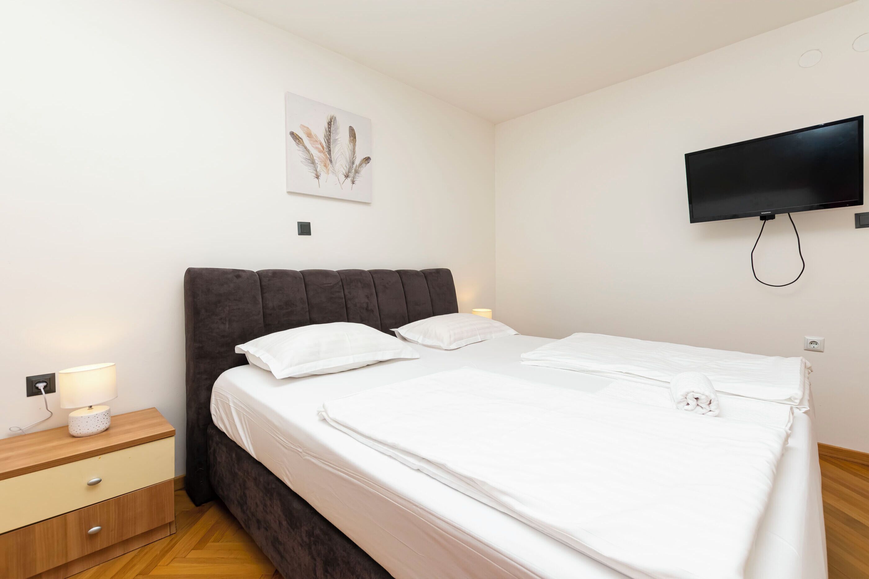 Ein ordentlich bezogenes Bett mit weißen Laken, einem dunklen Kopfteil und einem an der Wand montierten Fernseher.