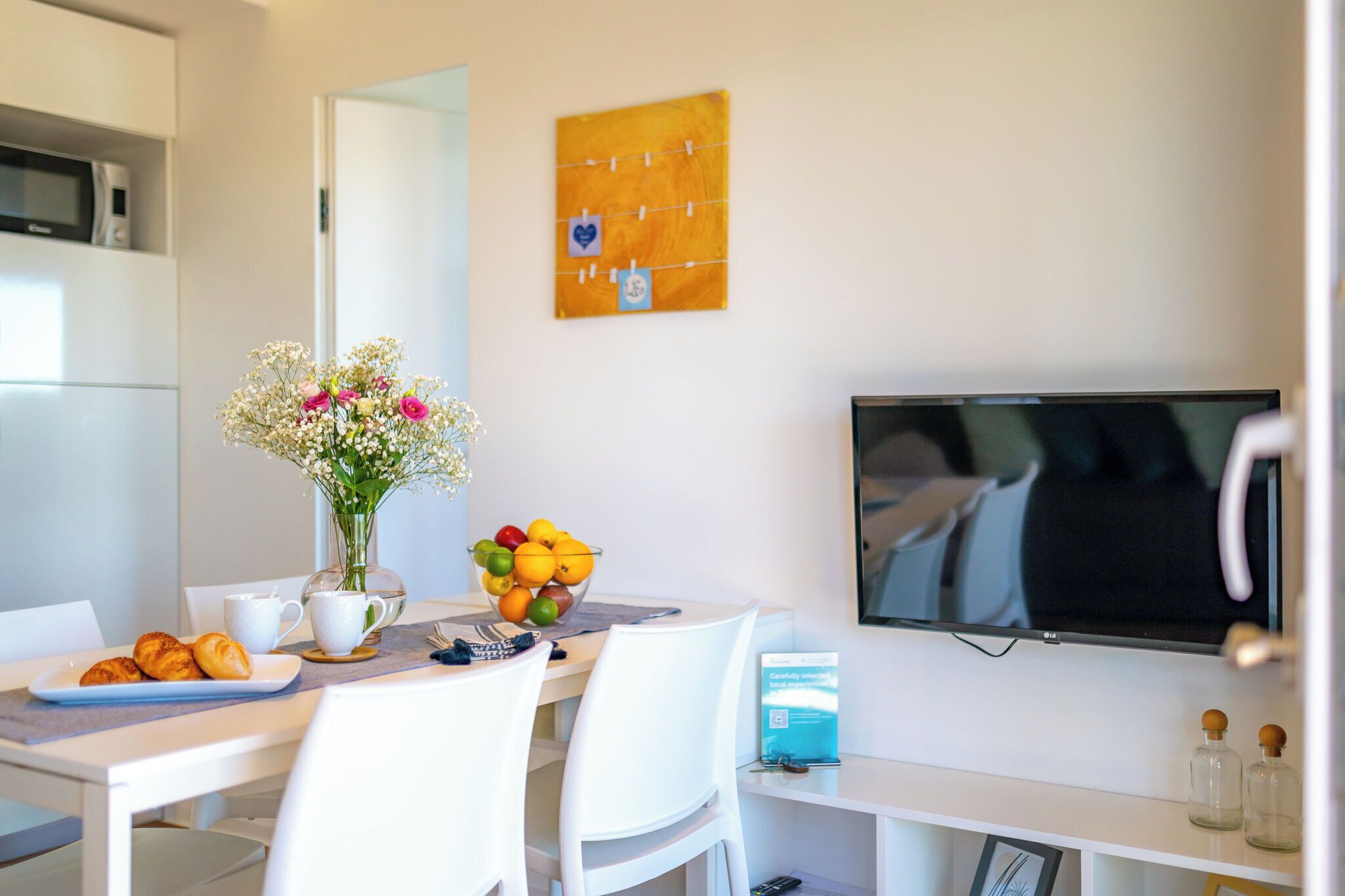 A modern dining area with a white table, chairs, a vase with flowers, a bowl of fruit, and a television mounted on the wall.