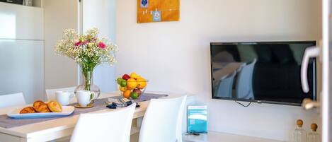 A modern dining area with a white table, chairs, a vase with flowers, a bowl of fruit, and a television mounted on the wall.