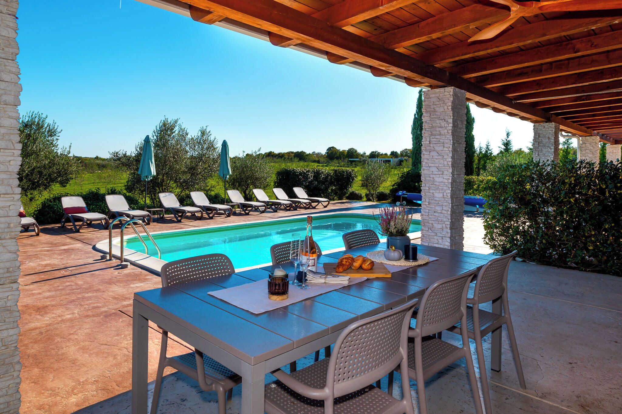 A poolside area with a covered patio, lounge chairs, and a table with drinks and pastries.