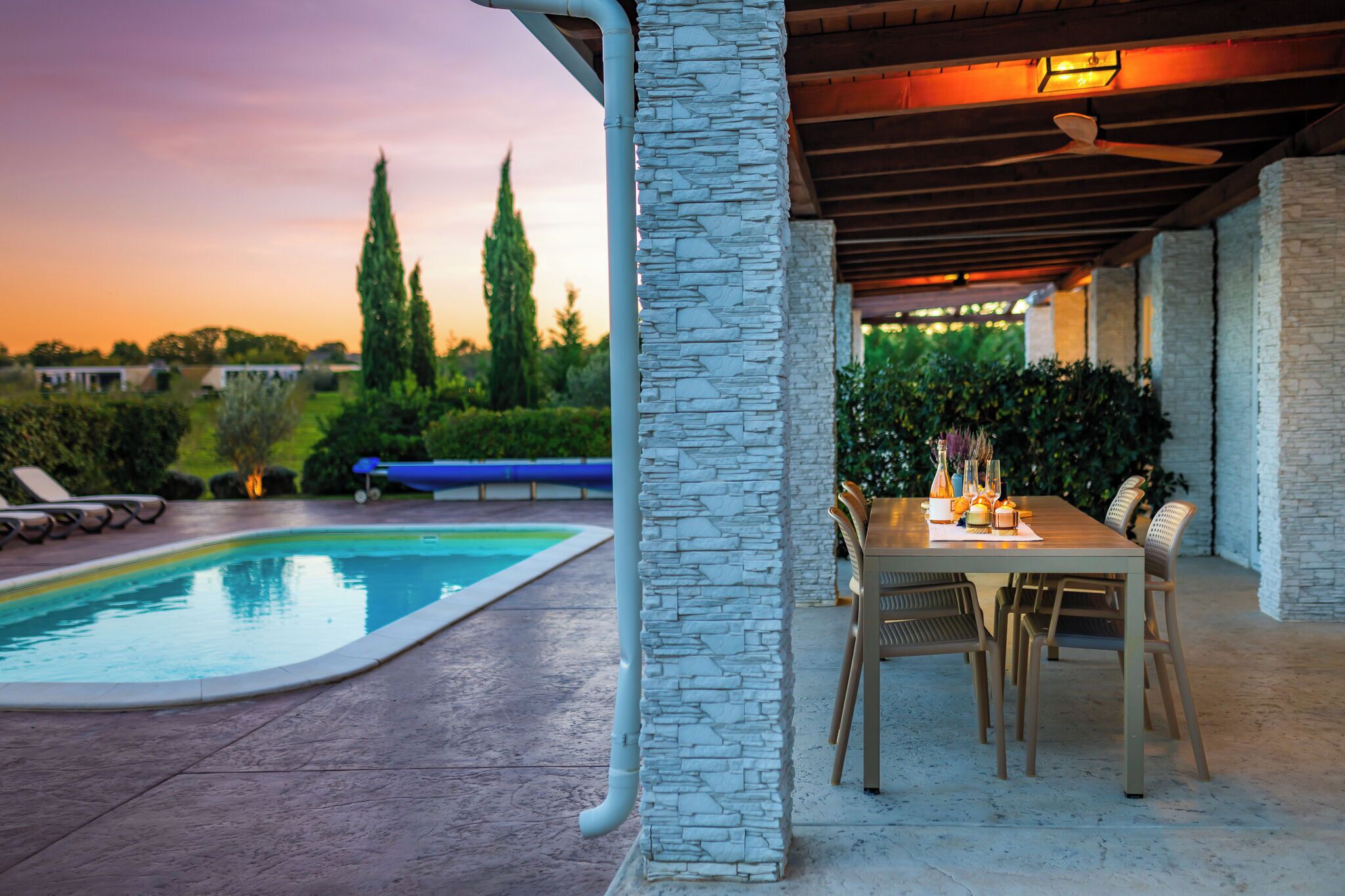Un espace aménagé au bord de la piscine, avec un pilier en pierre, une table et des chaises, et une vue sur le coucher du soleil.