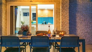 Un espace extérieur aménagé pour prendre les repas, avec une table dressée pour quatre personnes, entourée de chaises. Une bouteille et des verres sont posés sur la table, et une bougie est allumée.